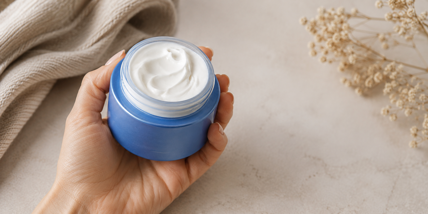 A hand holding an open blue jar of rich white face cream on a neutral surface with soft fabric and dried flowers in the background.