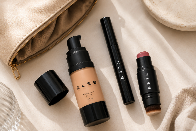 Four ELES cosmetics arranged on a beige fabric surface: a foundation bottle with its cap off, a black tube, a black stick, and a pinkish-brown lip/balm stick. A beige zip pouch and part of a dish are visible as styled props.