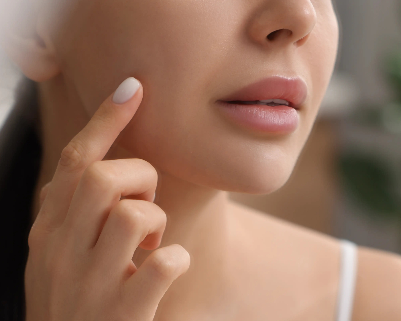 Close-up of a woman touching her cheek with her finger, showing smooth skin and soft pink lips, wearing a white strap top against a blurred indoor background.