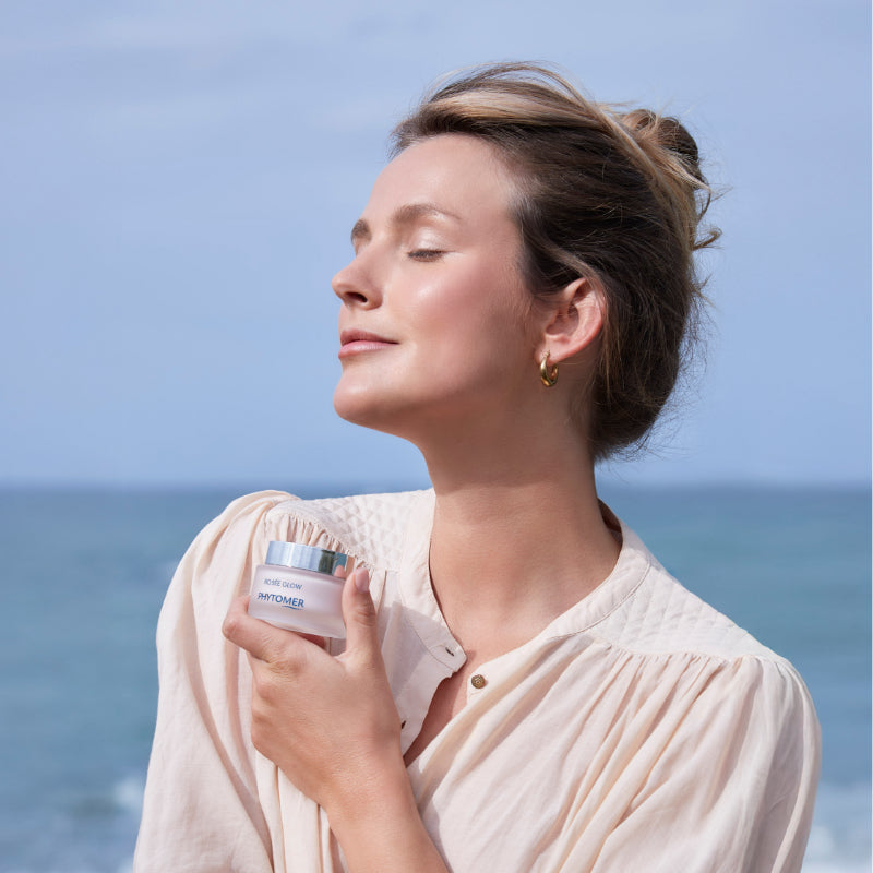 Woman holding Phytomer Rosee Glow - Vitamin Radiance Plumping Cream with ocean and sky in the background