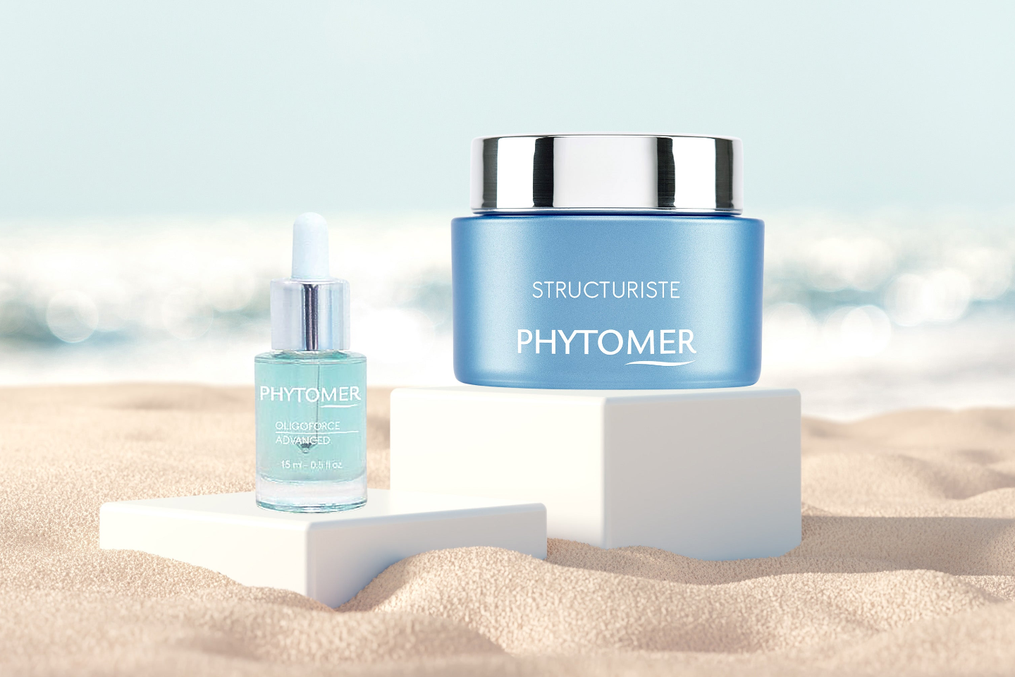 Two Phytomer skincare products, a blue cream jar and a light blue dropper bottle, styled on white pedestals in a sandy beach setting with a blurred ocean background.