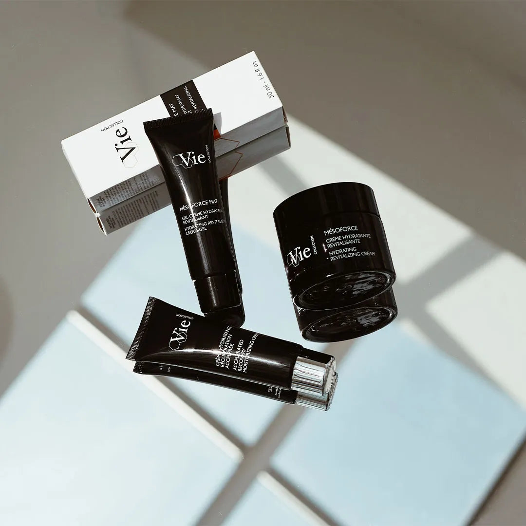 Vie! skincare products including a black jar, two black tubes, and a white box with black branding, arranged on a light gray surface with geometric shadows.