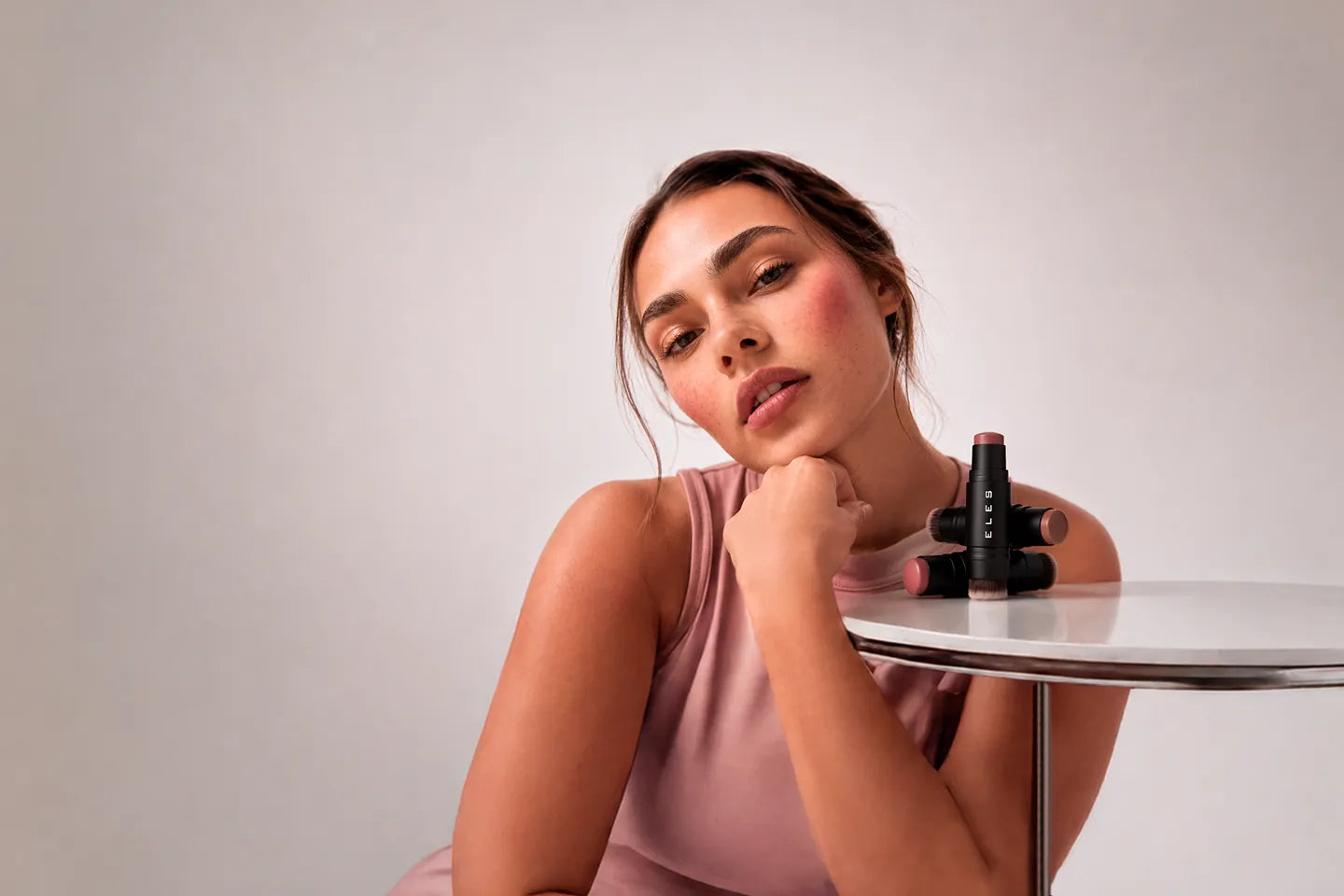 A woman in a dusty pink sleeveless top sits at a small round table, looking thoughtfully to the side with ELES Cheek & Lip Cream Blush