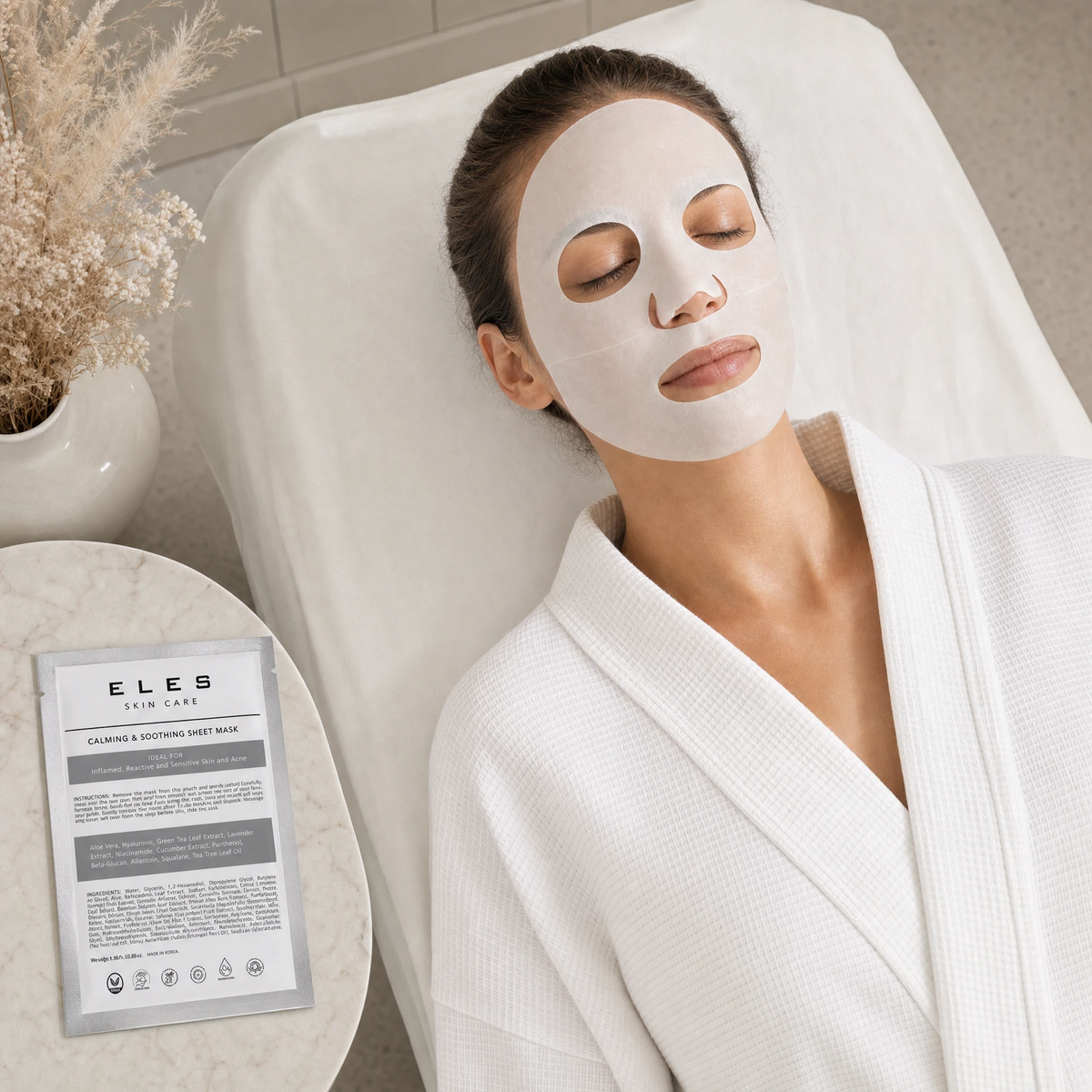 ELES Calming and Soothing Sheet Mask applied during a clinical skincare treatment, woman relaxing on treatment bed in white robe, silver foil sachet on marble side table, sensitive and reactive skin treatment