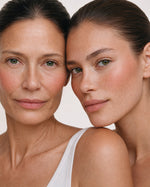 Two women with clean, natural skin posing closely together against a plain light background, wearing a white ribbed tank top.