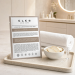 ELES Hydrating and Brightening Sheet Mask warm taupe foil sachet on a stone tray with ginseng root slices and a hyaluronate vial on a marble clinic bench inside a premium skin clinic treatment room