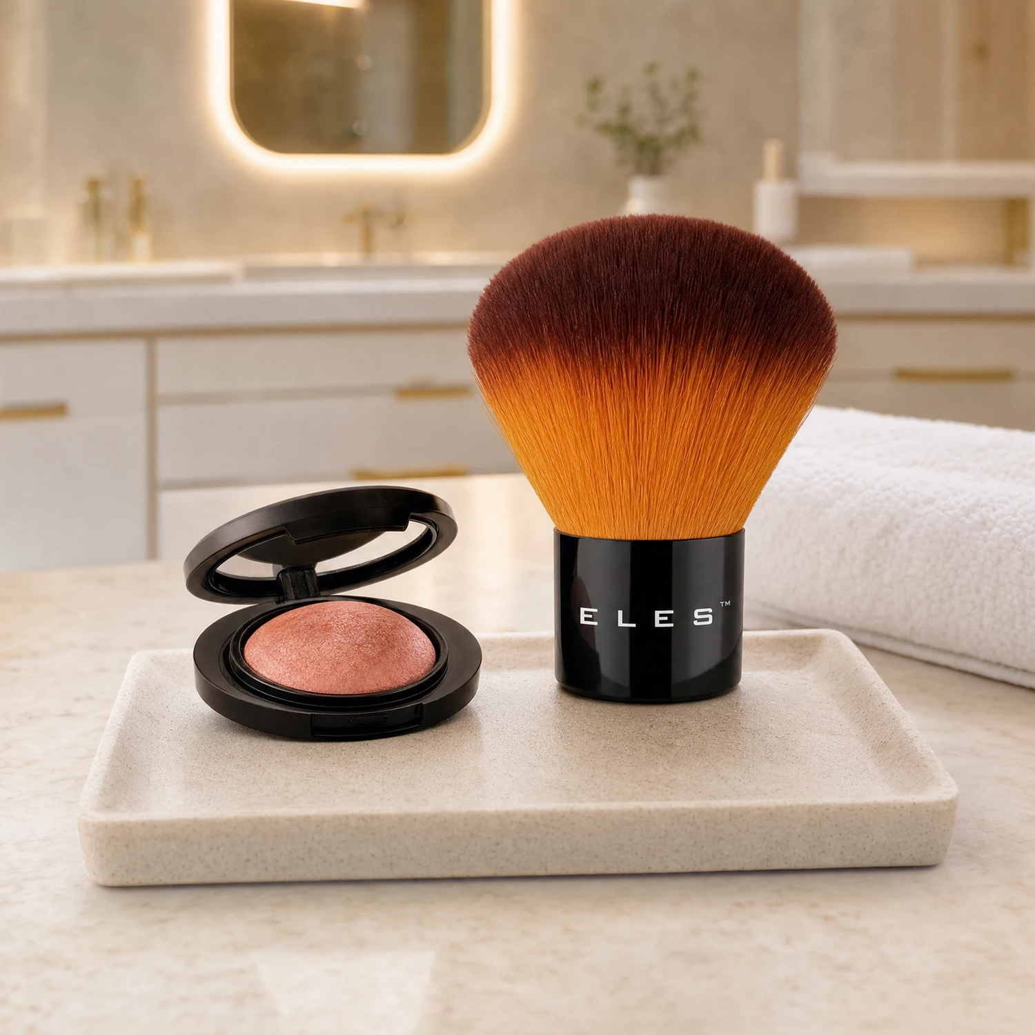 ELES Kabuki Brush with ombre amber bristles standing upright beside an open baked blush compact on a stone tray on a marble bench.