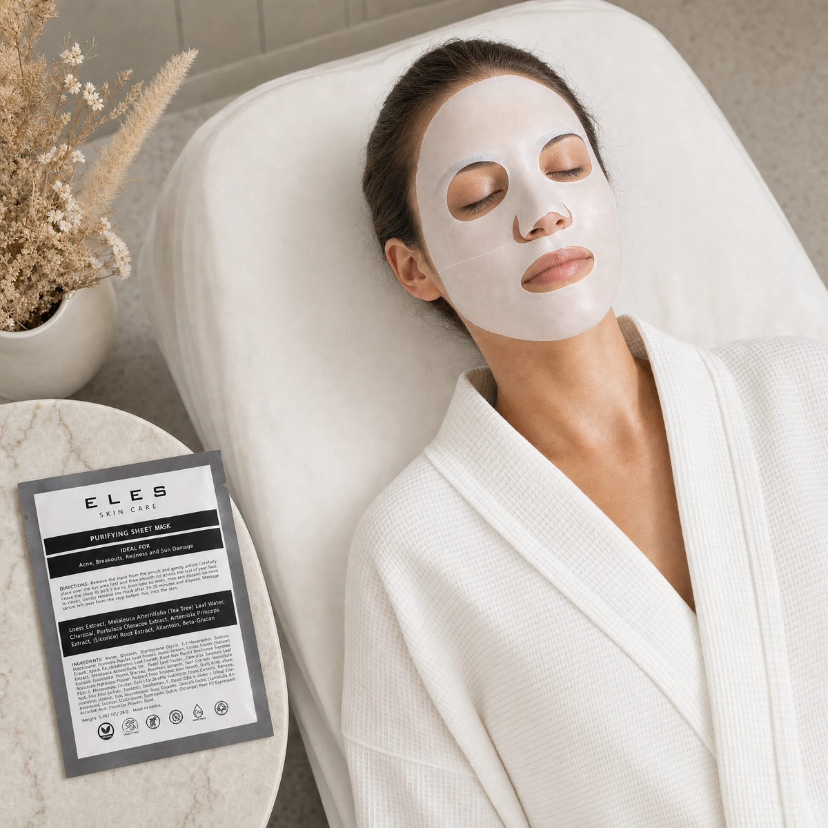 ELES Purifying Sheet Mask applied during a clinical skincare treatment, woman relaxing on treatment bed in white robe, charcoal grey foil sachet on marble side table, acne and breakout treatment