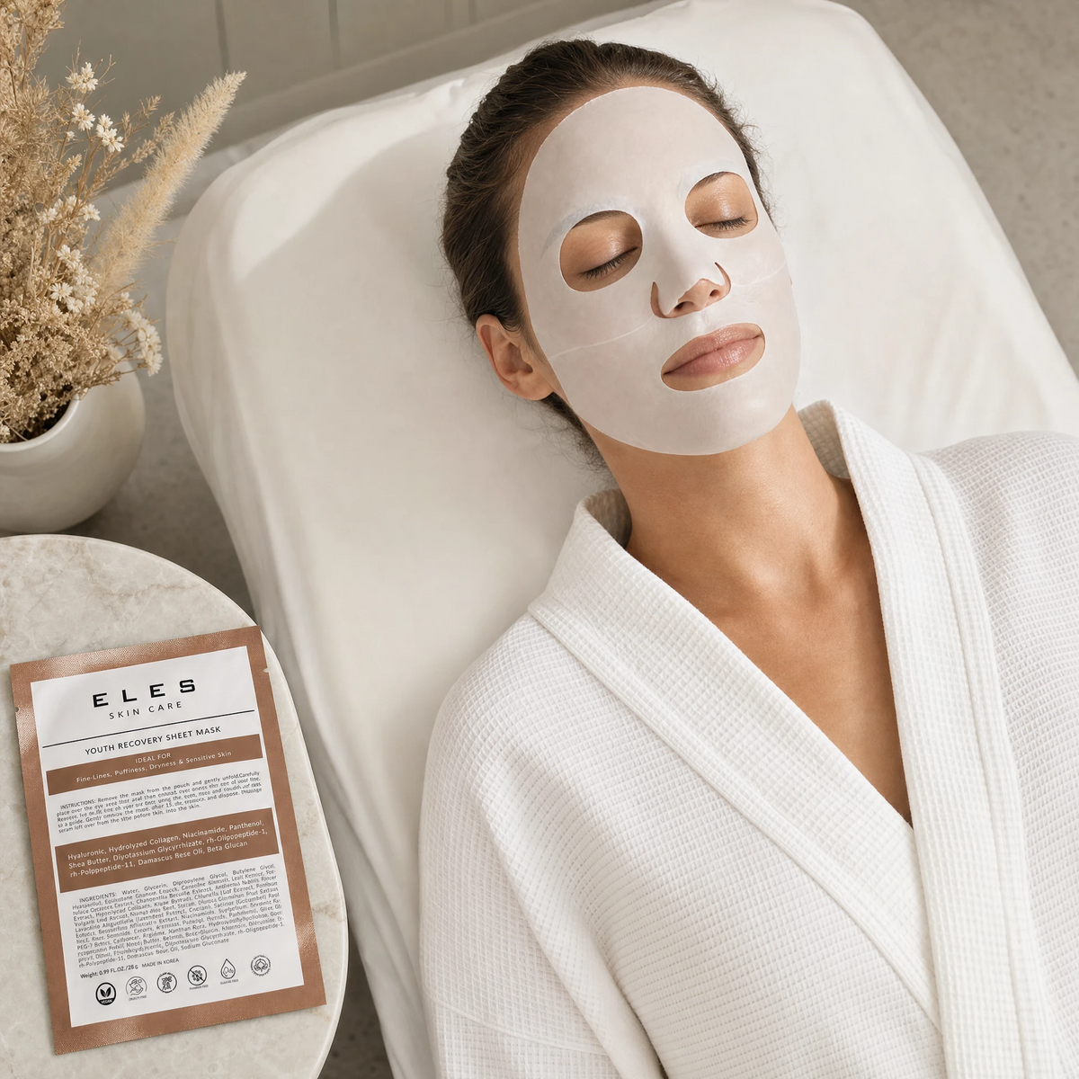 ELES Youth Recovery Sheet Mask applied during a clinical skincare treatment, woman relaxing on treatment bed in white robe, bronze foil sachet on marble side table
