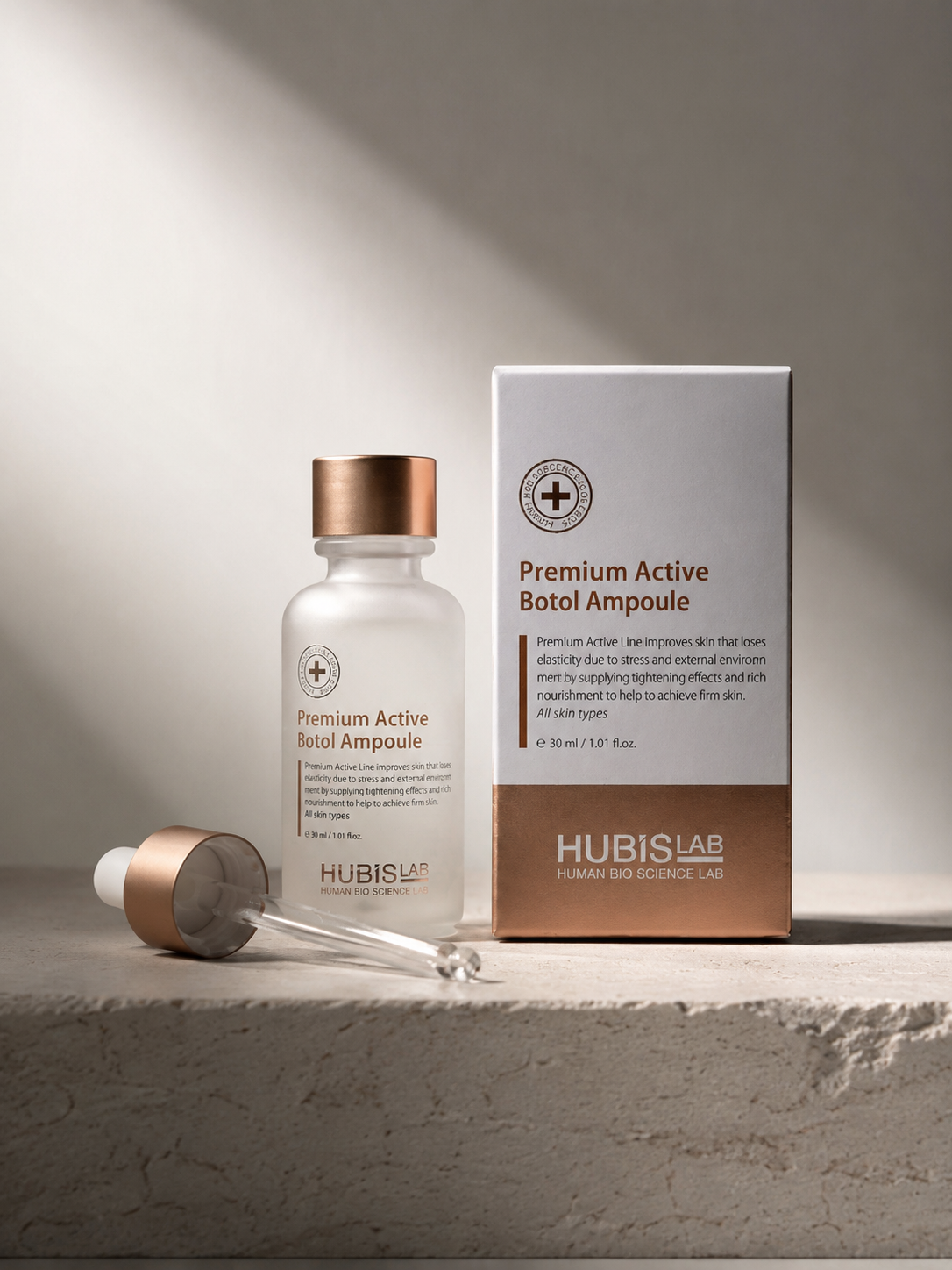HUBISLAB Premium Active Botol Ampoule set, featuring a glass bottle with a metallic rose gold cap, a separate glass dropper, and its white and rose gold product packaging box, arranged on a textured stone surface with dramatic diagonal lighting.
