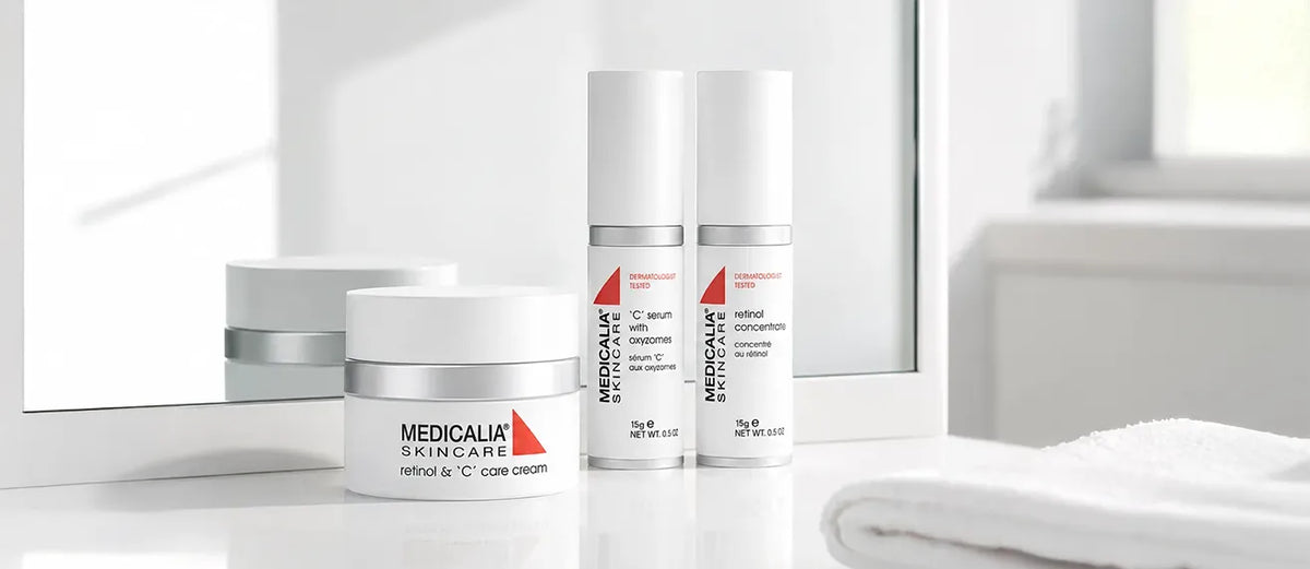 Three MEDICALIA SKINCARE skincare bottles and jars arranged on a glossy white surface in front of a softly blurred bathroom-like background with a mirror and folded towel.