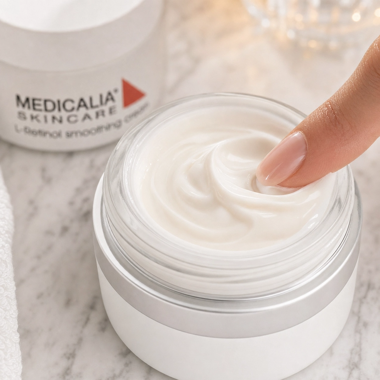 Close-up of Medicalia L-Retinol Smoothing Cream being scooped from an open jar showing the rich velvety cream texture with the branded product visible in the background on a marble surface