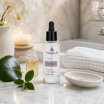 Naturys Anti-Age Multi-Peptide Serum 30ml clear glass dropper bottle on a white Carrara marble vanity with ceramic serum dish, vitamin C vial, fresh camellia leaf and white waffle linen cloth in a luxury candlelit spa bathroom with white orchids