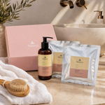 Naturys Body Box Cell anti-cellulite kit featuring pink box, amber pump bottle, and silver bandage sachets styled on a marble vanity with white towels and a dry body brush for Beauty Collective consumer retail.