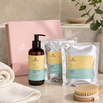 Naturys Body Box Dren firming kit featuring pink box, amber pump bottle, and silver bandage sachets styled on a marble vanity with white towels and a dry body brush for Beauty Collective consumer retail.