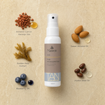 Naturys Tan Accelerating Spray Oil 100ml surrounded by labelled key ingredients including Annatto Carrot and Karanja Oils, Golden Kelp Extract, Blueberry Oil, Sweet Almond Oil and Jojoba Seed Oil on a warm golden travertine stone surface