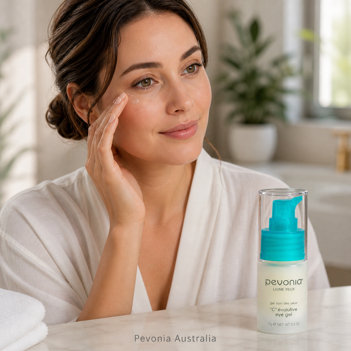 Woman applying Pevonia C Evolutive Eye Gel in a bright home bathroom setting with product displayed on countertop and subtle Pevonia Australia watermark at the bottom