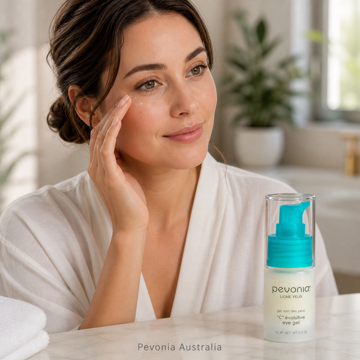 Woman applying Pevonia C Evolutive Eye Gel in a bright home bathroom setting with product displayed on countertop and subtle Pevonia Australia watermark at the bottom