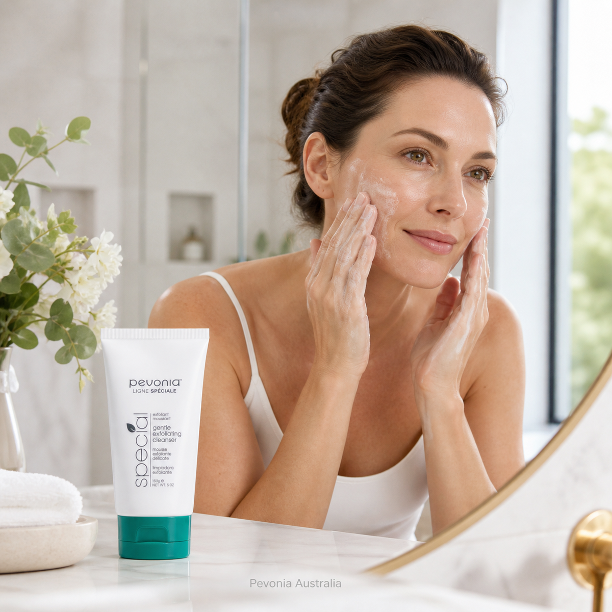 Woman using Pevonia Gentle Exfoliating Cleanser in a bright bathroom, showing a gentle daily exfoliating skincare routine with the cleanser tube displayed on the vanity.