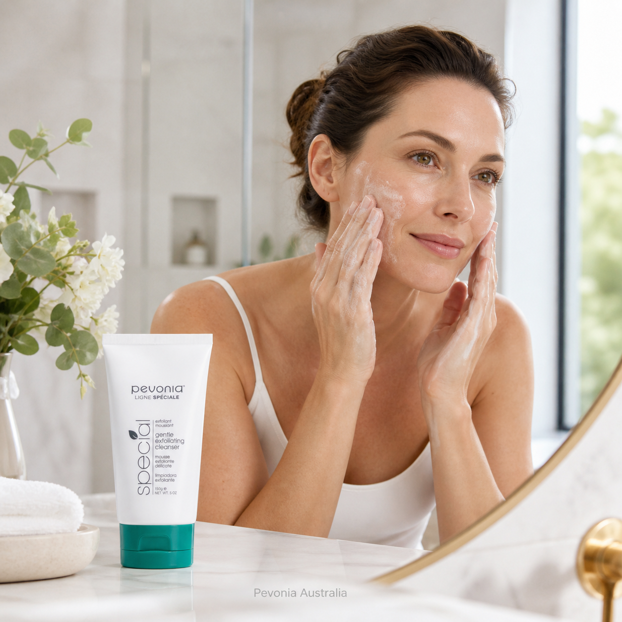 Woman using Pevonia Gentle Exfoliating Cleanser in a bright bathroom, showing a gentle daily exfoliating skincare routine with the cleanser tube displayed on the vanity.