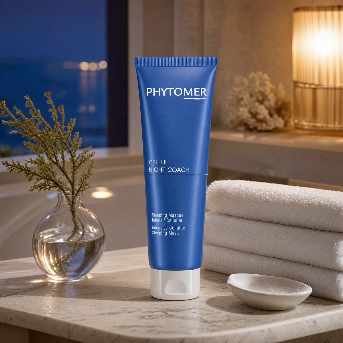 Phytomer Celluli Night Coach Intensive Cellulite Sleeping Mask 150ml on a marble bath surround with coastal botanical in glass vase, white coral, warm amber lamp and white towels, premium at-home evening body care setting