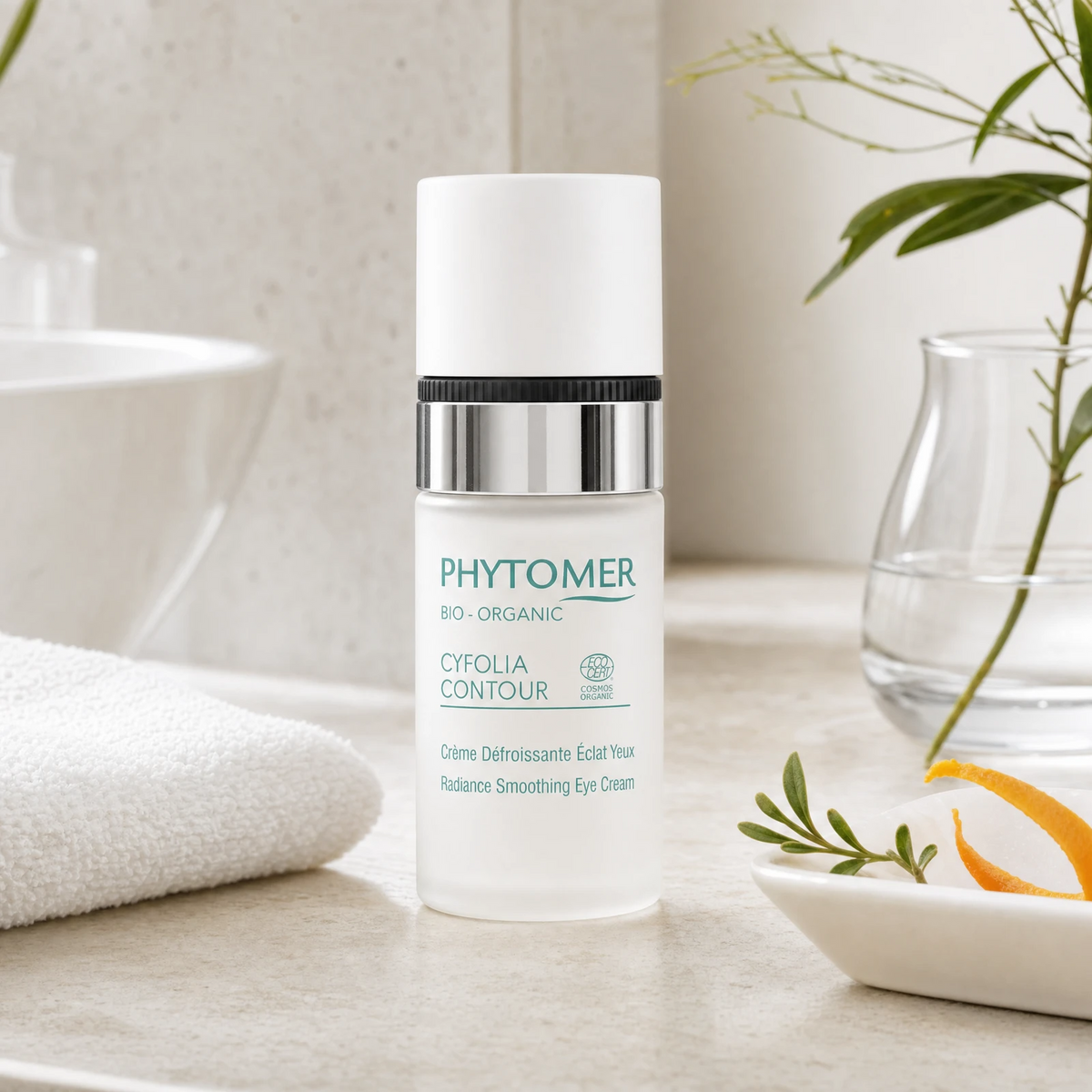 Phytomer Cyfolia Organic Radiance Smoothing Eye Cream 15ml on a marble bathroom counter with orange peel in ceramic dish, botanical in glass vase, rosemary sprig and white towels, premium at-home organic skincare setting