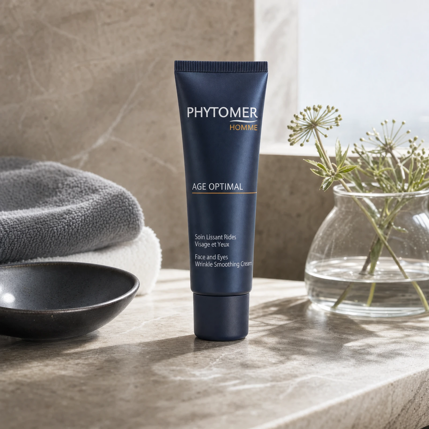 Phytomer Homme Age Optimal Face and Eyes Wrinkle Smoothing Cream 50ml on a dark marble grooming counter with dark ceramic dish, grey towel, glass vessel and sea fennel, premium men's grooming setting