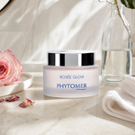 Phytomer Rosée Glow Vitamin Radiance Plumping Cream on a marble bathroom counter with fresh pink rose, scattered rose petals, glass carafe with water, white waffle towel and round mirror, premium at-home skincare setting
