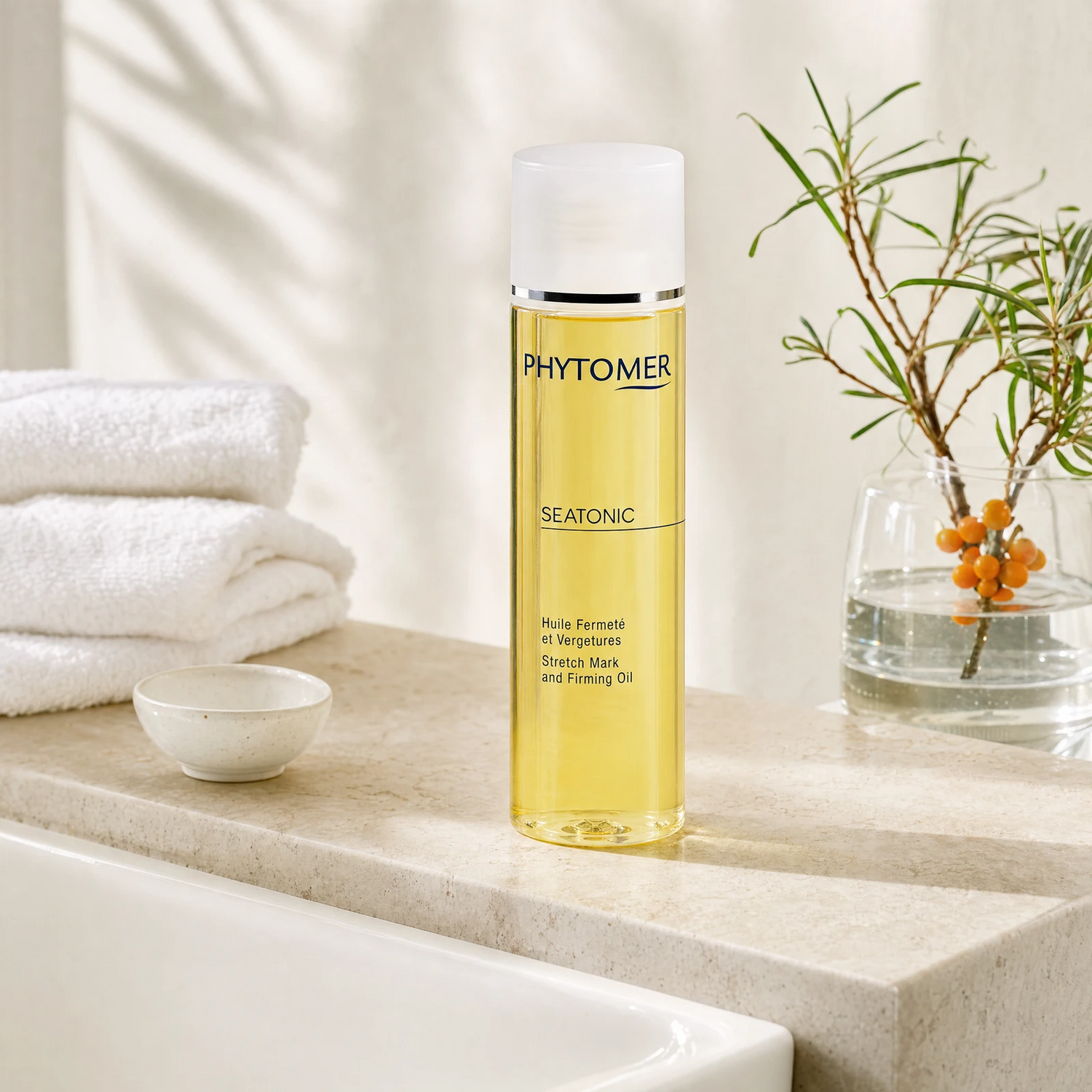 Phytomer Seatonic Stretch Mark and Firming Oil 125ml on a limestone bath surround with sea buckthorn sprig in glass vase, white ceramic bowl, white waffle towels and warm dappled light, premium at-home body care setting