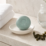 Phytomer Seaweed Soap 150g on a white ceramic soap dish on a marble bathroom vanity with white towels, glass vessel and dried seaweed, premium at-home bathroom setting