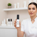 Woman holding a bottle of 'Natura' cream oil cleanser in a skincare setting.