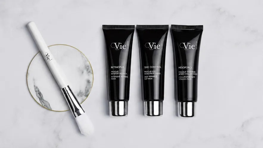 Three black Vie skincare tubes with silver caps are arranged on a white marble surface next to a white makeup brush resting on a round marble coaster.