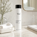 Vie Collection Instant Cleansing Milk 250ml on a marble bathroom counter with gypsophila in glass carafe, white ceramic tray with towel, gold round mirror and marble tile wall, premium at-home skincare setting