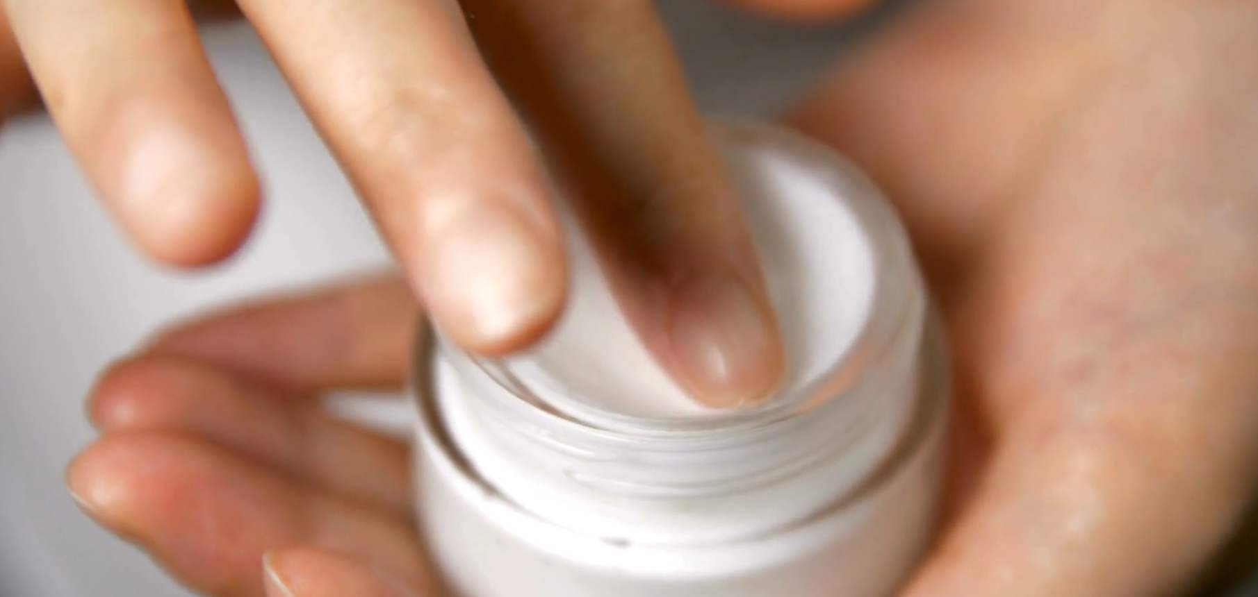 A close-up of a person's hand opening a small white cosmetic jar and touching the cream inside with their finger.